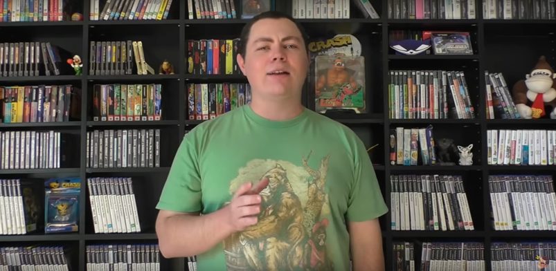 A man in a green shirt standing in a front of the black shelf with various things connected to games.