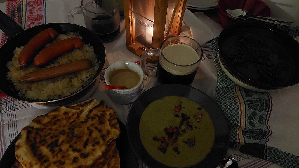 Foods on a table, over cloths. Foods include sausages, greenish soup, flatbreads, chocolate mousse, mustard and beer. In the middle of the table is a lantern, with candle burning inside.