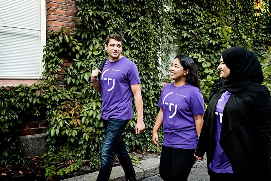 Kolme henkilöä pukeutuneena violettiin paitaan kävelee rakennuksen vierellä.