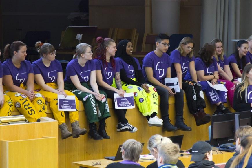 Joukko opiskelijoita luentosalin etuosassa. Kaikilla opiskelijoilla päällään violetti t-paita.