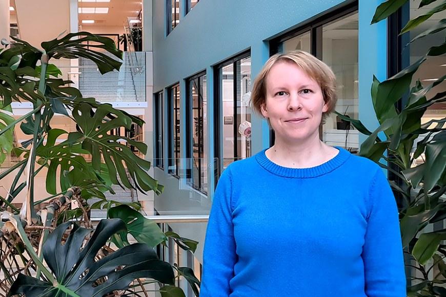 Mari Lehti-Polojärvi is wearing blue shirt and standing in front of big house plants. In the background is light blue wall with windows.