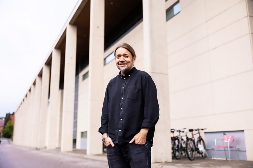 Markku-Juhani Saarinen stands with his hands in his pockets in front of a large light-colored building.