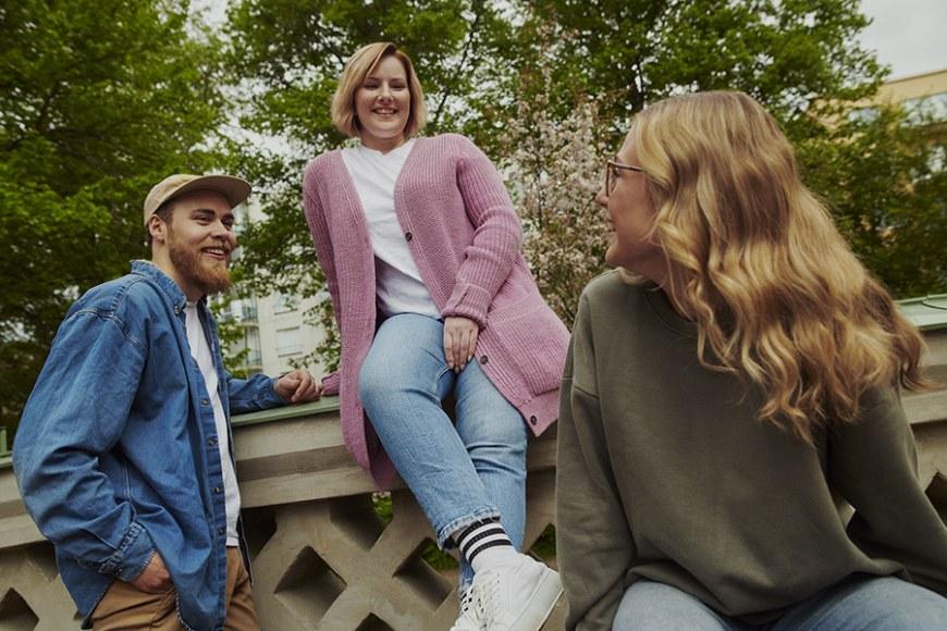 Nuori mies ja kaksi nuorta naista juttelevat hyväntuulisina puistomaisessa ympäristössä.