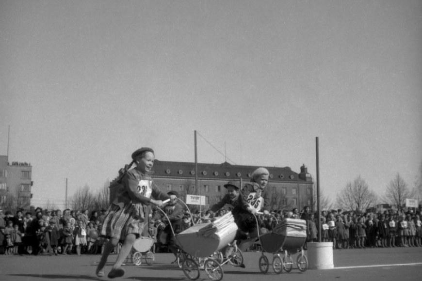 Mustavalkoinen kuva, jossa vanhassa kaupunkimaisemassa kaksi pientä lettipäistä tyttöä baskerit päässä kisaavat nukenvaunujen työnnössä. Kisa on meneillään ja toisen tytön vaunut ovat keikahtamaisillaan.
