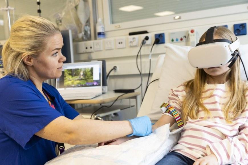 Lapsi istuu sairaalasängyssä VR-lasit päässään. Naishoitaja pitää häntä kädestä ja on suorittamassa pistoshoitotoimenpidettä lapselle. Taustalla kannettava tietokone, jonka näytöllä näkyy VR-sovelluksen kuvitus. 