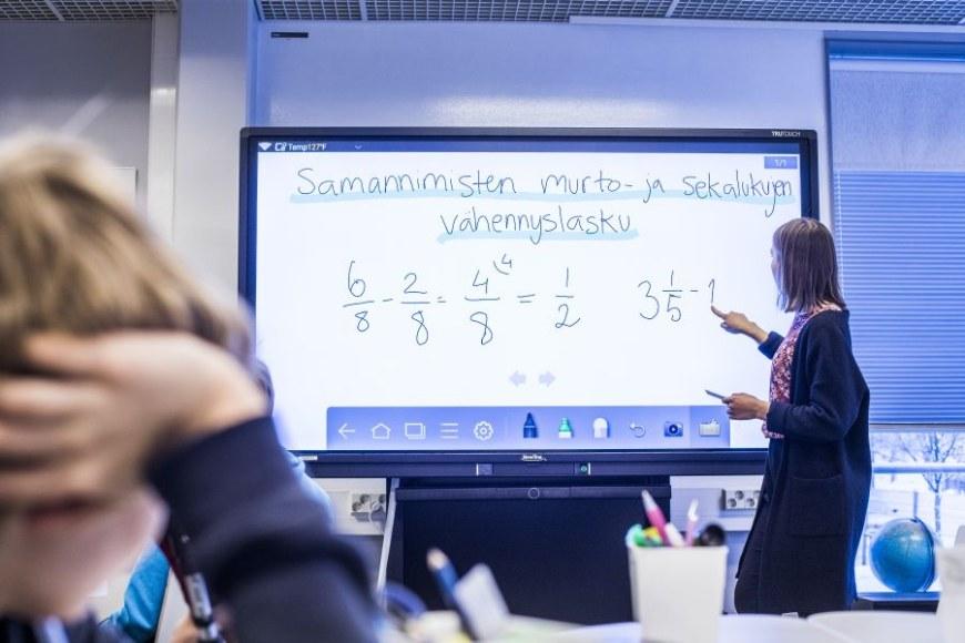 Opettaja opettaa matematiikkaa. Hän osoittaa digitaalisella taululla olevaa laskutehtävää.
