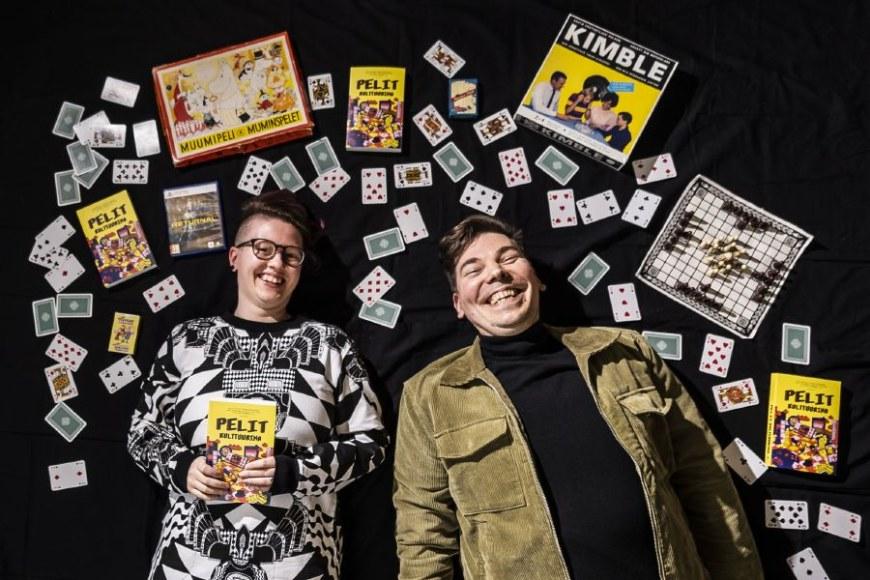 Kuvassa kaksi kirjan toimittajista makaavat lattialla nauraen. Heidän ympärillään on erilaisia suomalaisia pelejä, pelikortteja ja Pelit kulttuurina -kirjoja.
