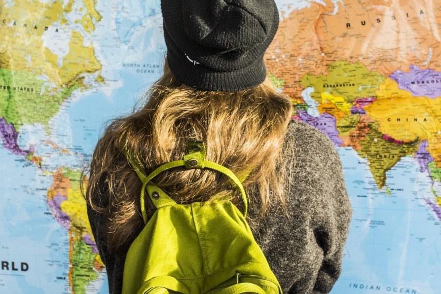 Neulepaitaan ja myssyyn pukeutunut, vihreää reppua selässään kantava, opiskelija katsoo maailman karttaa. 