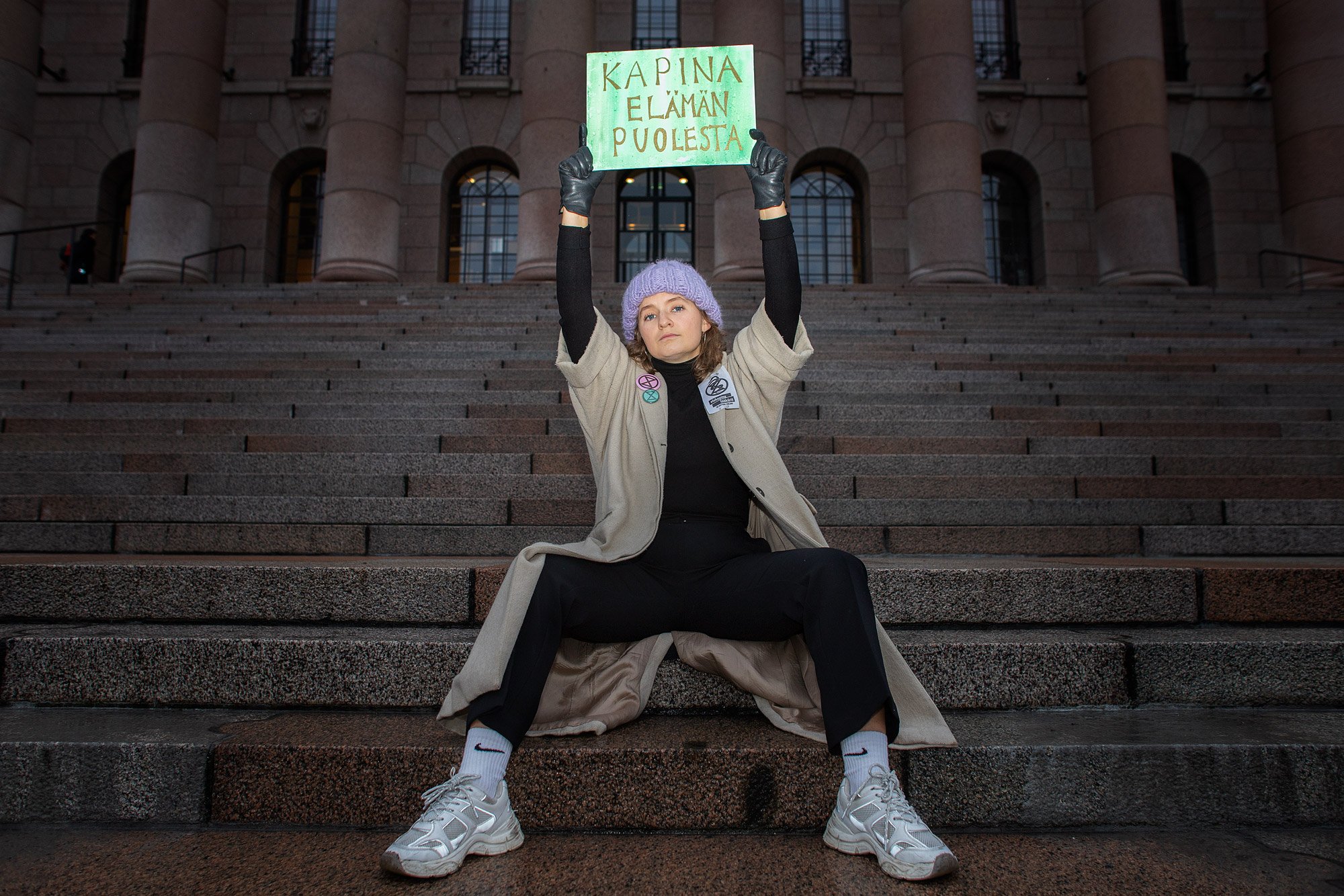 Nainen keskellä kuvaa. Istuu eduskuntatalon portailla Helsingissä. Pitää käsissään mielenosoitus kylttiä.