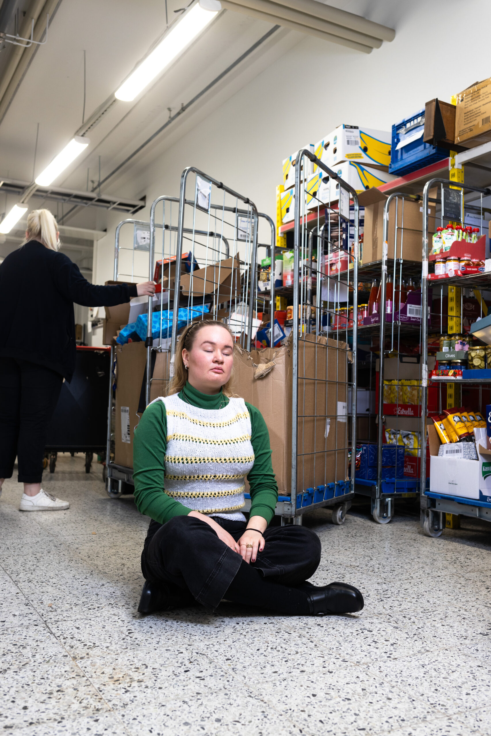 Minna Arpiainen istuu ruokakaupan varaston lattialla. Takana työntekijä tekee töitään.