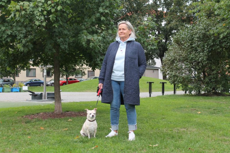 Minna Kauppila seisoo Sorsapuistossa. Hänellä on hihnan päässä pieni sekarotuinen Milli-koira, joka istuu hänen vieressään.