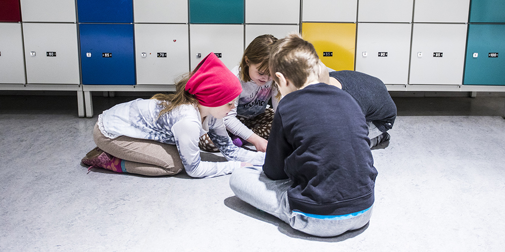 Neljä alakoulun pikkuoppilasta ovat käytävän lattialla piirissä istuen tekemässä yhteistä tehtävää.
