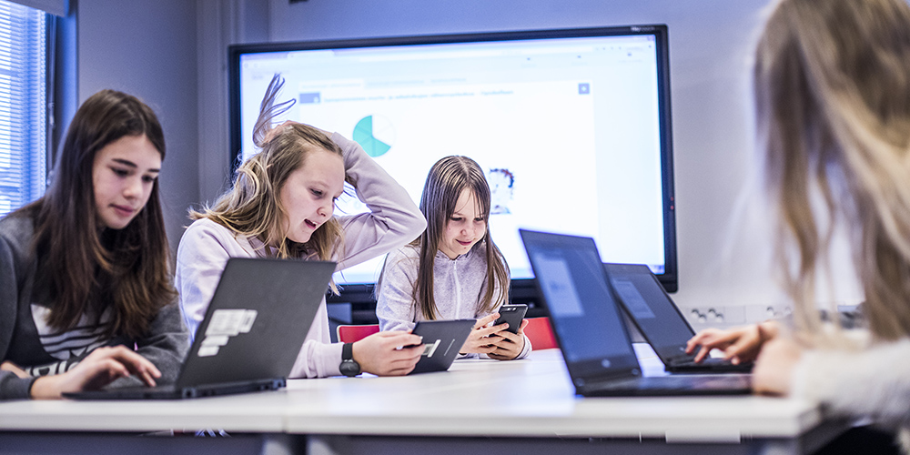 Kuvassa kuudesluokkalaisen tytöt istuvat vierekkäin erilaisia päätelaitteita käyttäen. Yhdellä on tablet-laite, yhdellä puhelin ja kolmella kannettavat tietokoneet.