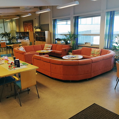 Kuva Norssin henkilöstöhuoneesta, jossa on suuret oranssit sohvat, pöytiä ja irtotuoleja sekä kirjahyllyjä.
