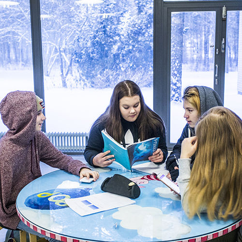 Kuvassa on neljä oppilasta pöydän ääressä koulun aulassa välitunnilla. Taustalla on suuri ikkuna, josta näkyy talvinen metsikkö.
