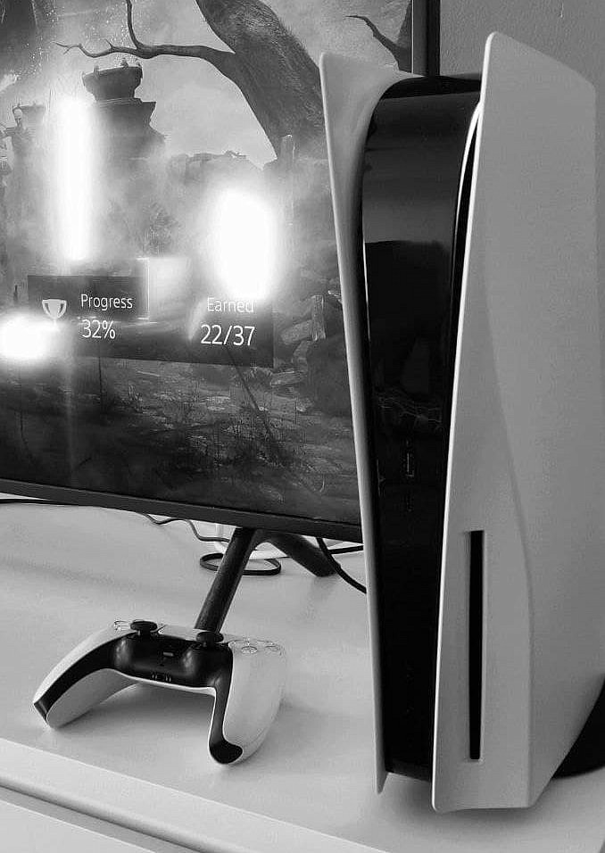 The PlayStation 5 is at the forefront with the controller next to it. Behind is some gameplay on a TV screen.