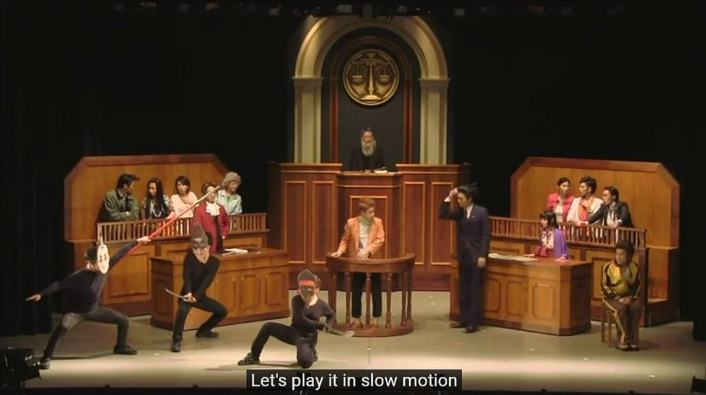 Courtroom in session on a stage, with actors acting out security camera footage and being controlled by the defence attorney holding the remote