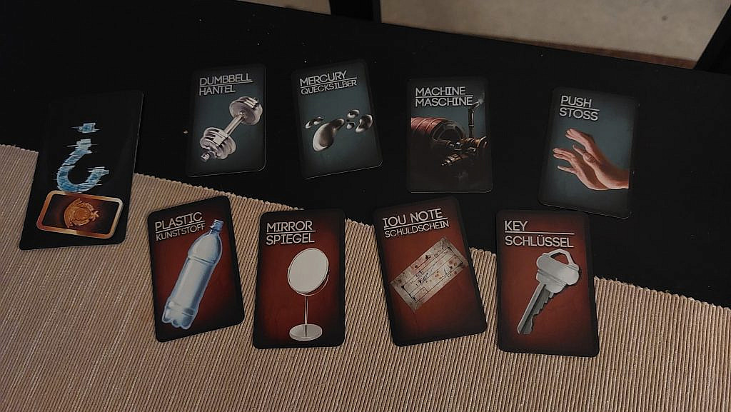 A set of 8 cards face-up displaying various murder weapons and items. A face-down card with a question mark, and a small cardboard token on top of the card.