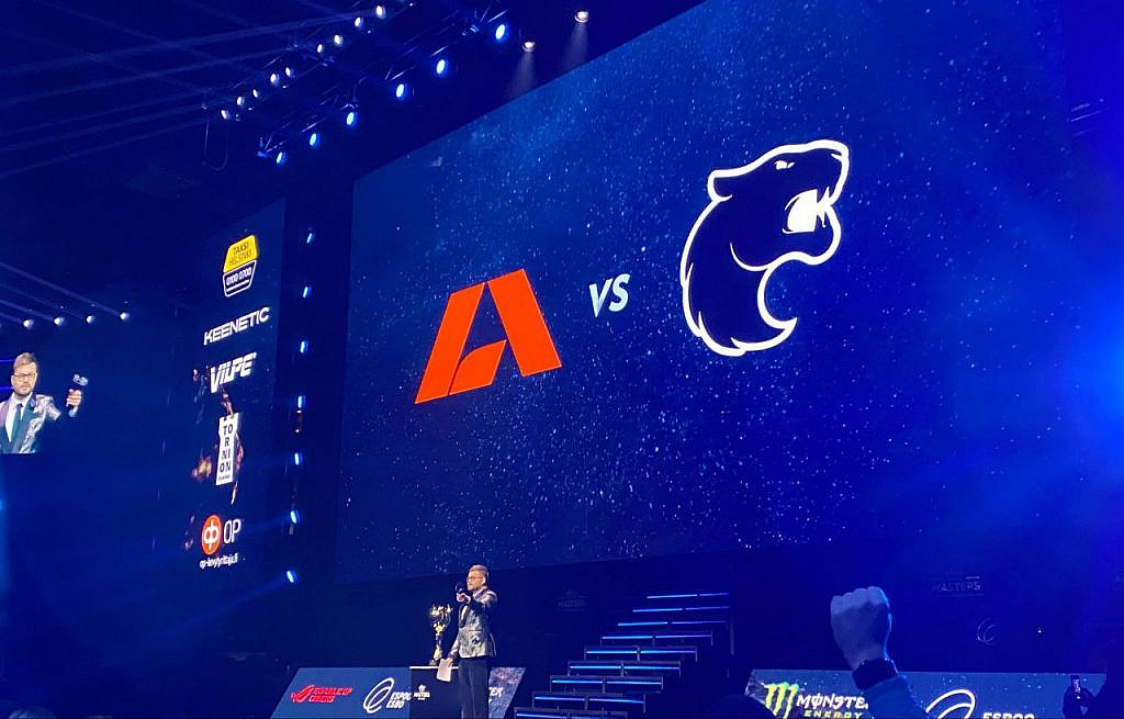 Host on stage with the final duo's logos in the back