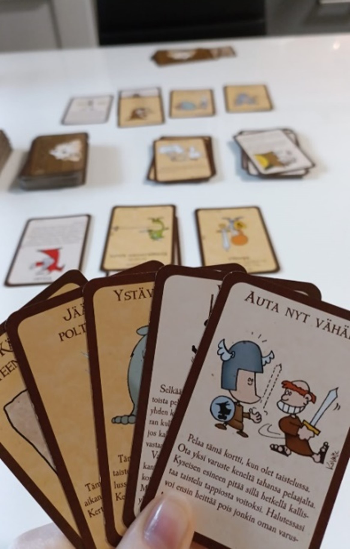 Five brown cards with funny pictures in the hand of the player and many cards on the table in the background.