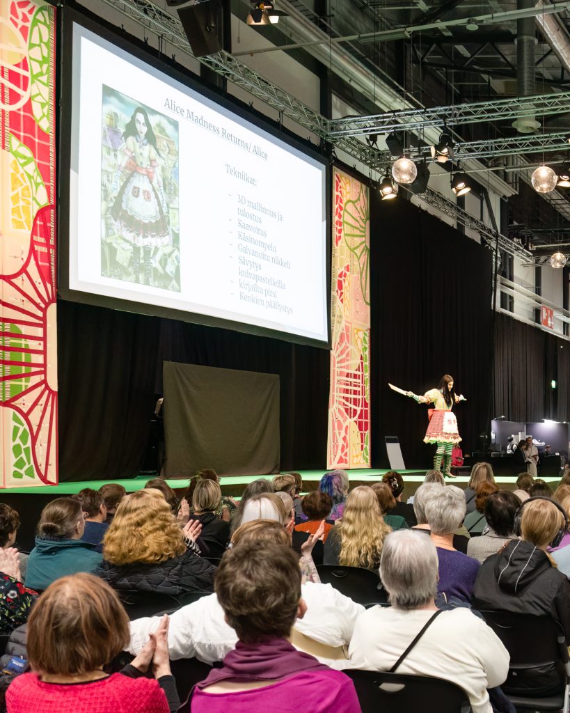 Cosplayer on the main stage, dressed as Alice from the videogame Alice: Madness Returns. The audience in the foreground is clapping.