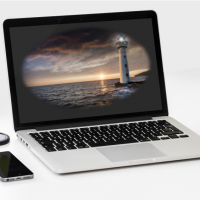 Kuvassa on tietokone, jonka näytöllä on merimaisema ja majakka. Koneen vieressä on mobiililaite ja omena.