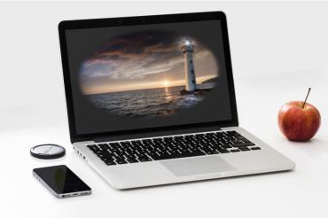Kuvassa on tietokone, jonka näytöllä on merimaisema ja majakka. Koneen vieressä on mobiililaite ja omena.