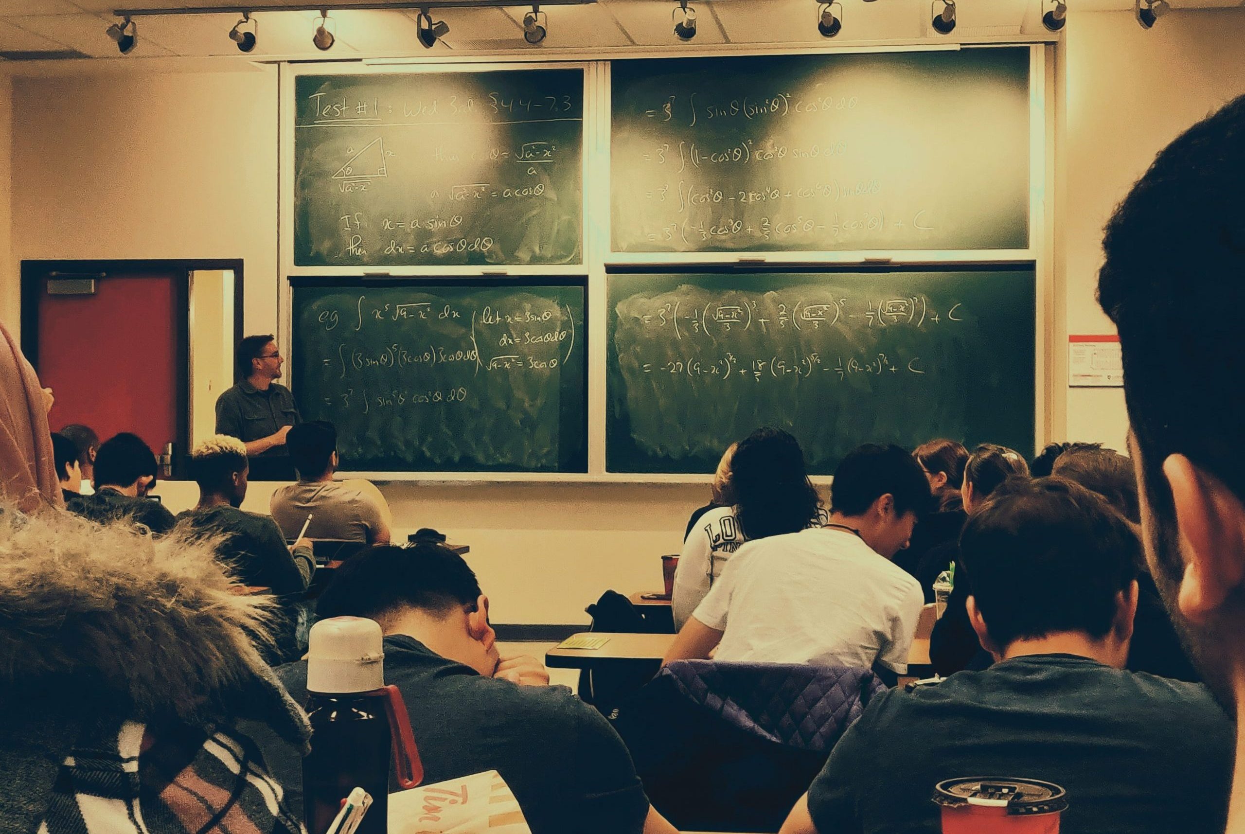 Opettaja liitutaulun luona, opiskelijat seuraavat matemaattisten aineiden opetusta.